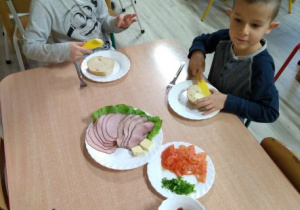 Chłopcy smarują masłem bułeczki. Na stoliku stoją talerze z wędliną, warzywami, salaterka z dżemem